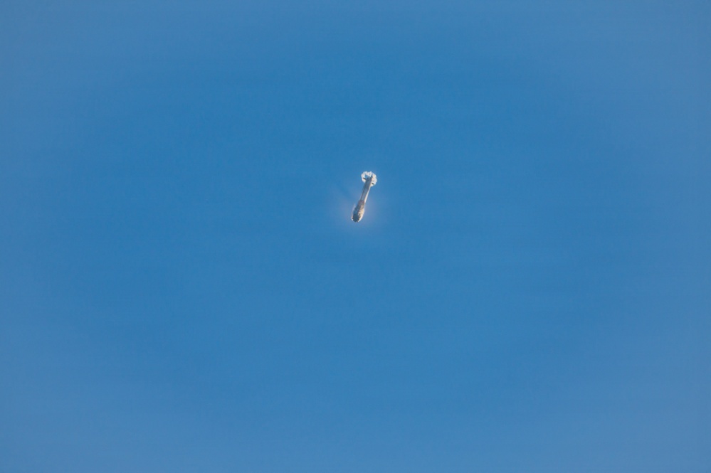 The image shows a small, elongated white and dark object photographed against a clear, uniformly blue sky. The object appears to be at high altitude, making precise identification difficult due to distance. It has a cylindrical or oblong shape with what appears to be a bright reflective area at one end.