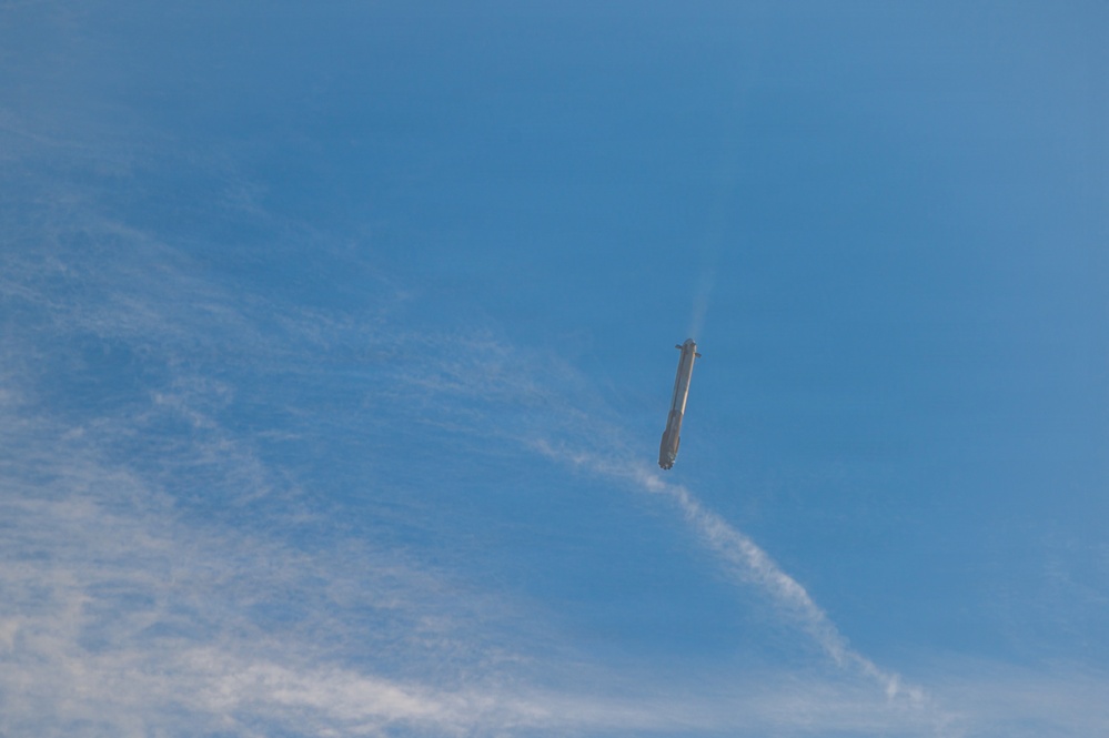 A rocket or large missile is captured mid-flight against a blue sky with scattered cirrus clouds. The vehicle appears to be oriented nearly vertically, with a visible exhaust plume or vapor trail emanating from its upper end, suggesting it may be in a descent or reentry phase. The image was taken from a ground-level perspective looking upward.
