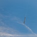 A rocket or large missile is captured mid-flight against a blue sky with scattered cirrus clouds. The vehicle appears to be oriented nearly vertically, with a visible exhaust plume or vapor trail emanating from its upper end, suggesting it may be in a descent or reentry phase. The image was taken from a ground-level perspective looking upward.