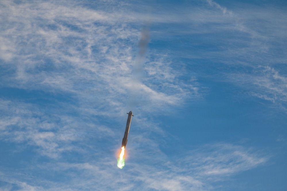 A large rocket is captured mid-flight against a blue sky with wispy clouds, with its engines actively firing and producing a bright orange and green flame plume. The rocket appears to be ascending at a near-vertical angle, with a dark smoke trail visible above it. The image appears to depict a SpaceX Starship or similar large launch vehicle during a test flight.