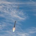 A large rocket is captured mid-flight against a blue sky with wispy clouds, with its engines actively firing and producing a bright orange and green flame plume. The rocket appears to be ascending at a near-vertical angle, with a dark smoke trail visible above it. The image appears to depict a SpaceX Starship or similar large launch vehicle during a test flight.