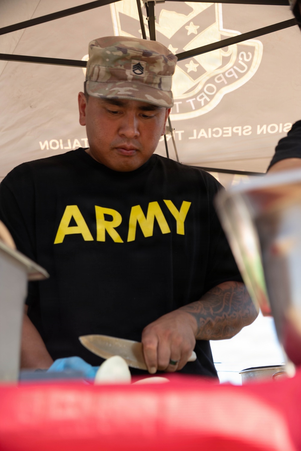 A U.S. Army soldier wearing a black 'ARMY' PT shirt and a multicam patrol cap with a Staff Sergeant rank insignia is cutting food at what appears to be an outdoor cooking or food service event. He is working under a tent canopy that displays text referencing a 'Mission Special Troops Battalion' or similar unit. The setting suggests a community event, cookout, or military outreach activity.