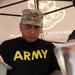 A U.S. Army soldier wearing a black 'ARMY' PT shirt and a multicam patrol cap with a Staff Sergeant rank insignia is cutting food at what appears to be an outdoor cooking or food service event. He is working under a tent canopy that displays text referencing a 'Mission Special Troops Battalion' or similar unit. The setting suggests a community event, cookout, or military outreach activity.