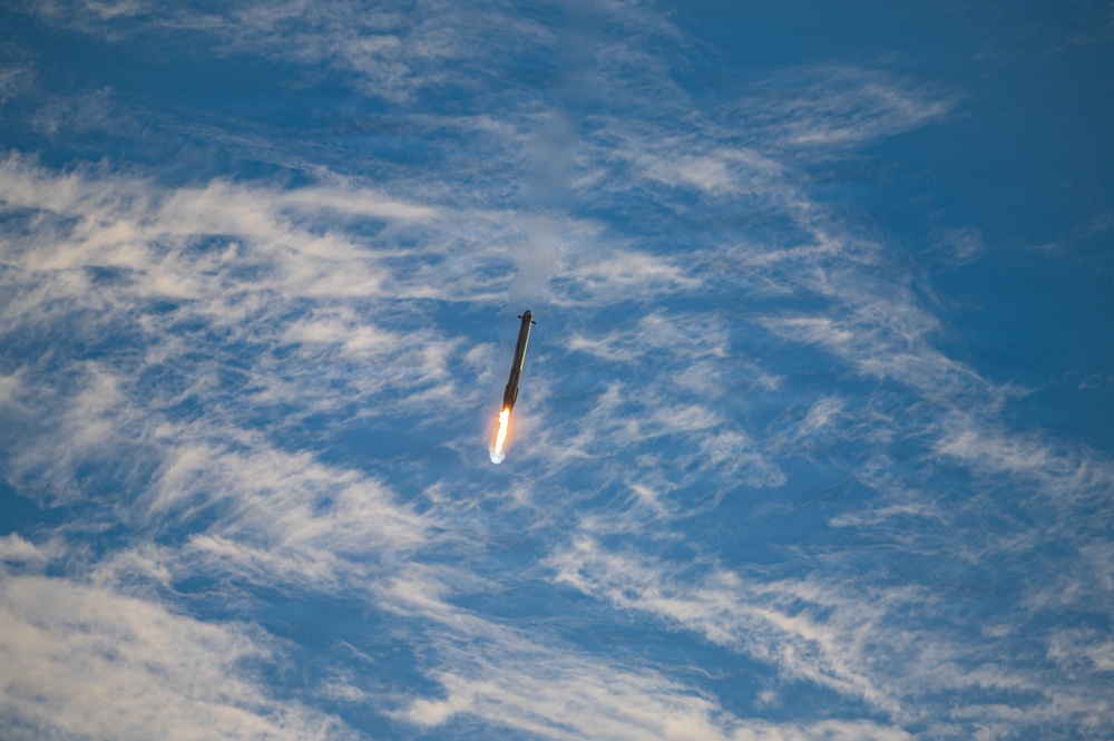A rocket is captured mid-flight against a blue sky with scattered white clouds. The rocket appears to be ascending vertically with a visible engine plume of orange and white flame at its base. The image was likely taken from a distance, possibly from an aircraft or elevated vantage point.