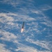 A rocket is captured mid-flight against a blue sky with scattered white clouds. The rocket appears to be ascending vertically with a visible engine plume of orange and white flame at its base. The image was likely taken from a distance, possibly from an aircraft or elevated vantage point.