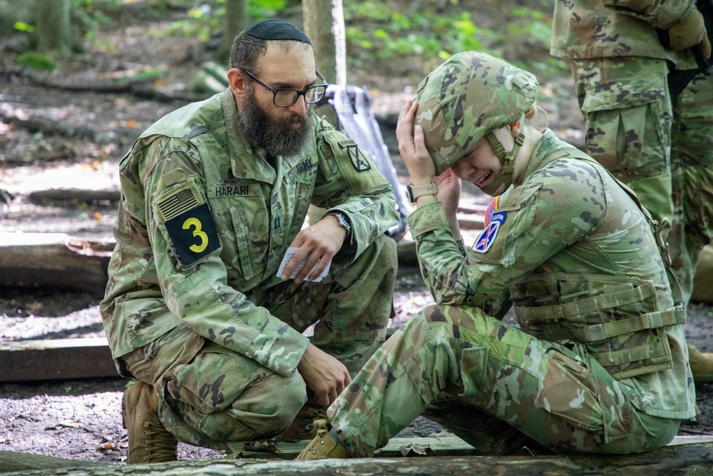 A U.S. Army chaplain or officer named HARARI, wearing a kippah and bearing a '3' unit patch, crouches beside a distressed soldier who is seated on the ground with their hand on their helmet. The interaction appears to be a pastoral counseling or welfare check moment in a wooded outdoor training environment. A third soldier is partially visible in the background.