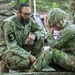A U.S. Army chaplain or officer named HARARI, wearing a kippah and bearing a '3' unit patch, crouches beside a distressed soldier who is seated on the ground with their hand on their helmet. The interaction appears to be a pastoral counseling or welfare check moment in a wooded outdoor training environment. A third soldier is partially visible in the background.