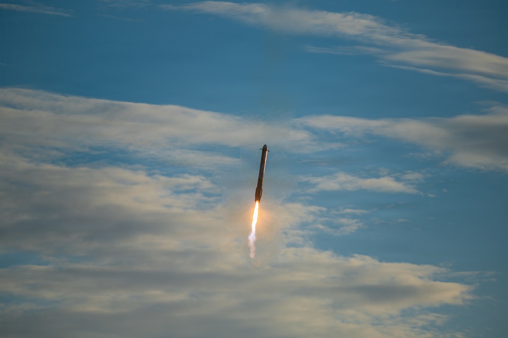 A rocket is captured mid-flight ascending vertically through a partly cloudy blue sky. Bright orange and white engine exhaust flames and a white smoke trail are clearly visible beneath the vehicle. The rocket appears dark-colored and slender, photographed from a ground-level perspective looking upward.