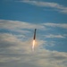 A rocket is captured mid-flight ascending vertically through a partly cloudy blue sky. Bright orange and white engine exhaust flames and a white smoke trail are clearly visible beneath the vehicle. The rocket appears dark-colored and slender, photographed from a ground-level perspective looking upward.