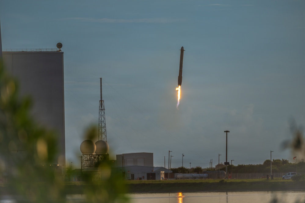 The image captures a rocket booster in a near-vertical descent with engine firing, consistent with a controlled landing attempt. The scene is set at dusk near a launch facility with industrial buildings, a water body in the foreground, and a lattice communications tower visible. The rocket appears to be in the final moments of a propulsive landing maneuver.