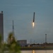 The image captures a rocket booster in a near-vertical descent with engine firing, consistent with a controlled landing attempt. The scene is set at dusk near a launch facility with industrial buildings, a water body in the foreground, and a lattice communications tower visible. The rocket appears to be in the final moments of a propulsive landing maneuver.