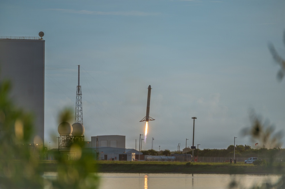 A rocket booster is captured mid-descent during a landing attempt at what appears to be a coastal launch facility, with its engine firing a bright flame downward as it approaches the ground. The scene is photographed across a body of water at dusk or dawn, with facility infrastructure visible in the background. The rocket's landing legs are deployed, indicating this is a controlled propulsive landing maneuver.