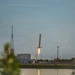 A rocket booster is captured mid-descent during a landing attempt at what appears to be a coastal launch facility, with its engine firing a bright flame downward as it approaches the ground. The scene is photographed across a body of water at dusk or dawn, with facility infrastructure visible in the background. The rocket's landing legs are deployed, indicating this is a controlled propulsive landing maneuver.