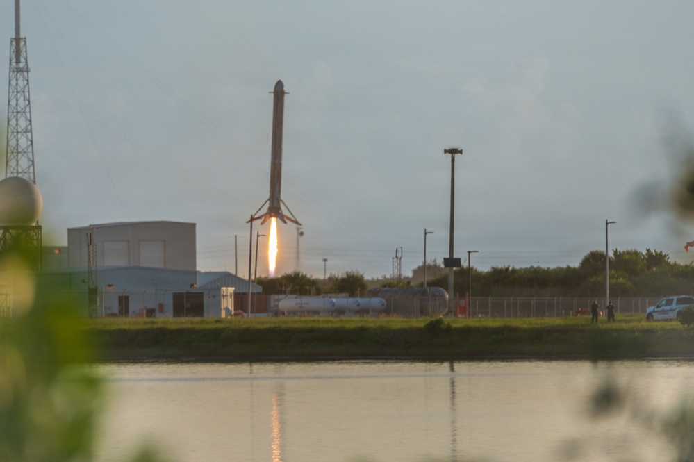 A SpaceX Falcon 9 rocket booster is captured in the moment of landing, with its landing legs deployed and engine firing a bright flame to slow its descent. The scene is photographed across a body of water at what appears to be dusk or dawn, with launch facility infrastructure visible in the background. Several observers and a vehicle are visible near a perimeter fence to the right of the frame.