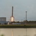 A SpaceX Falcon 9 rocket booster is captured in the moment of landing, with its landing legs deployed and engine firing a bright flame to slow its descent. The scene is photographed across a body of water at what appears to be dusk or dawn, with launch facility infrastructure visible in the background. Several observers and a vehicle are visible near a perimeter fence to the right of the frame.