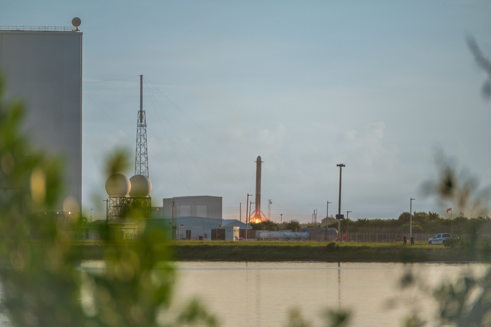 A rocket booster is visible standing upright on a landing pad at what appears to be a SpaceX launch facility, with a bright engine fire or landing burn visible at its base. The scene is photographed across a body of water at dusk or dawn, with facility buildings, a lattice tower, and spherical storage tanks visible in the background. Foreground vegetation and the reflective water surface frame the distant industrial scene.
