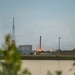 A rocket booster is visible standing upright on a landing pad at what appears to be a SpaceX launch facility, with a bright engine fire or landing burn visible at its base. The scene is photographed across a body of water at dusk or dawn, with facility buildings, a lattice tower, and spherical storage tanks visible in the background. Foreground vegetation and the reflective water surface frame the distant industrial scene.