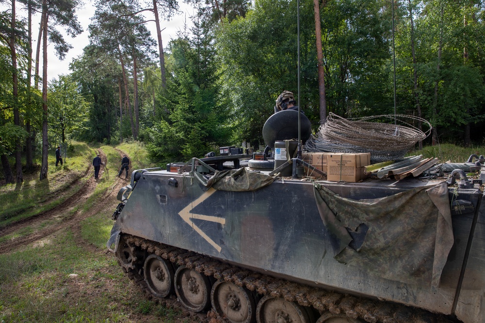 A military armored personnel carrier (APC), identifiable as an M113 variant, is positioned on a dirt track at the edge of a forested area. The vehicle is loaded with equipment including coiled wire, cardboard boxes, and camouflage netting, with one soldier visible in the hatch. Several personnel are visible in the background on the dirt path.