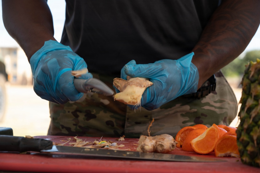 A person wearing blue nitrile gloves and military camouflage pants is peeling or preparing fresh ginger root on a red cutting board outdoors. Several fruits and ingredients are visible on the surface, suggesting food preparation in a field or outdoor setting. The close-up framing emphasizes the hands and food preparation activity.