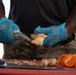 A person wearing blue nitrile gloves and military camouflage pants is peeling or preparing fresh ginger root on a red cutting board outdoors. Several fruits and ingredients are visible on the surface, suggesting food preparation in a field or outdoor setting. The close-up framing emphasizes the hands and food preparation activity.