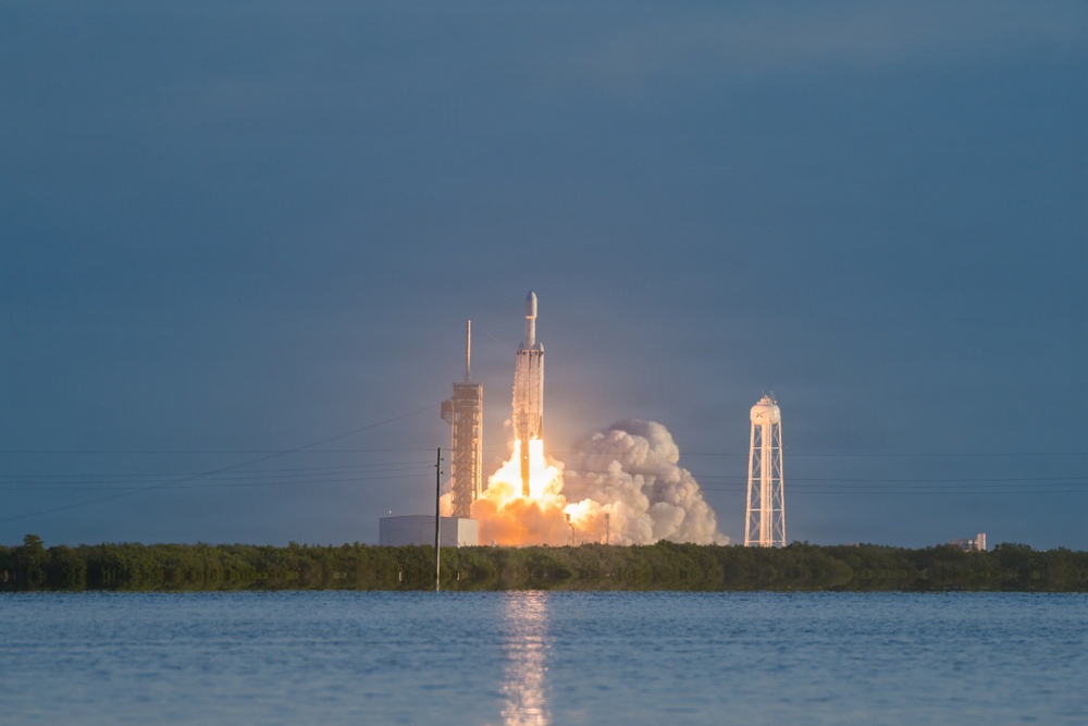 A large rocket is captured at the moment of launch from a coastal launch pad, with massive plumes of fire and white smoke billowing outward. The scene is photographed from across a body of water, reflecting the launch light on its surface. The launch infrastructure, including a service tower and a water tower, is visible against a cloudy, overcast sky.