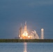 A large rocket is captured at the moment of launch from a coastal launch pad, with massive plumes of fire and white smoke billowing outward. The scene is photographed from across a body of water, reflecting the launch light on its surface. The launch infrastructure, including a service tower and a water tower, is visible against a cloudy, overcast sky.