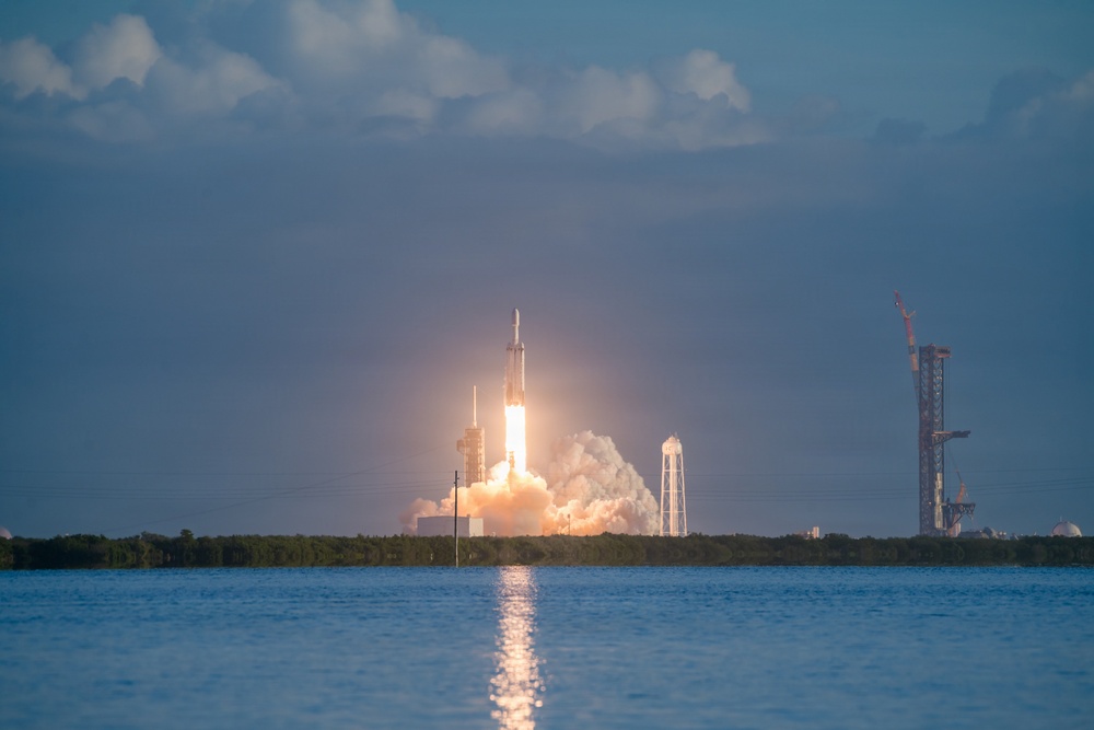 A large rocket is captured mid-launch from a coastal launch facility, producing a massive plume of white smoke and bright orange flame at its base. The rocket appears to be a heavy-lift vehicle with a distinctive triple-core configuration, consistent with a Falcon Heavy-type design. The launch is photographed across a body of water, with a secondary launch structure and construction crane visible in the background.
