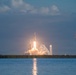 A large rocket is captured mid-launch from a coastal launch facility, producing a massive plume of white smoke and bright orange flame at its base. The rocket appears to be a heavy-lift vehicle with a distinctive triple-core configuration, consistent with a Falcon Heavy-type design. The launch is photographed across a body of water, with a secondary launch structure and construction crane visible in the background.
