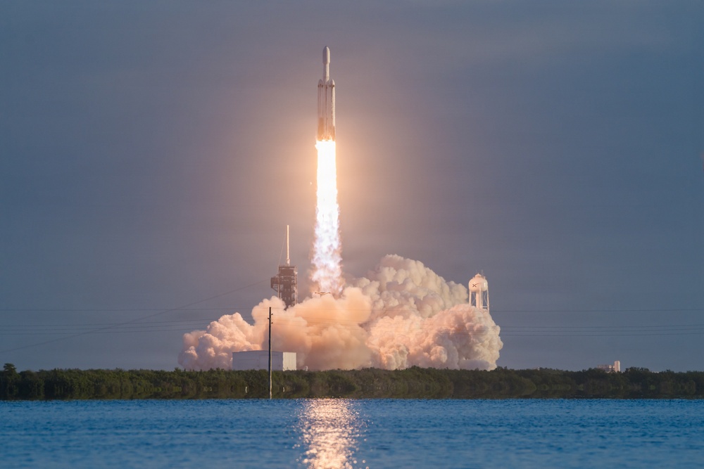 A large rocket is captured mid-launch, ascending vertically with a bright plume of fire and billowing clouds of exhaust smoke. The launch appears to take place at a coastal facility, with a body of water visible in the foreground and a treeline in the background. The rocket's distinctive triple-core configuration is visible, consistent with a heavy-lift launch vehicle.