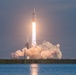 A large rocket is captured mid-launch, ascending vertically with a bright plume of fire and billowing clouds of exhaust smoke. The launch appears to take place at a coastal facility, with a body of water visible in the foreground and a treeline in the background. The rocket's distinctive triple-core configuration is visible, consistent with a heavy-lift launch vehicle.