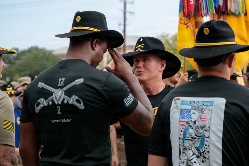 The image shows a military ceremony or gathering involving U.S. Army Cavalry soldiers wearing distinctive black campaign (Stetson) hats with gold trim. A senior officer, identifiable by a general's star insignia on his hat, appears to be adjusting or presenting a hat to another soldier. The setting includes unit colors/guidons and multiple soldiers in black unit t-shirts in the background.