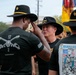 The image shows a military ceremony or gathering involving U.S. Army Cavalry soldiers wearing distinctive black campaign (Stetson) hats with gold trim. A senior officer, identifiable by a general's star insignia on his hat, appears to be adjusting or presenting a hat to another soldier. The setting includes unit colors/guidons and multiple soldiers in black unit t-shirts in the background.