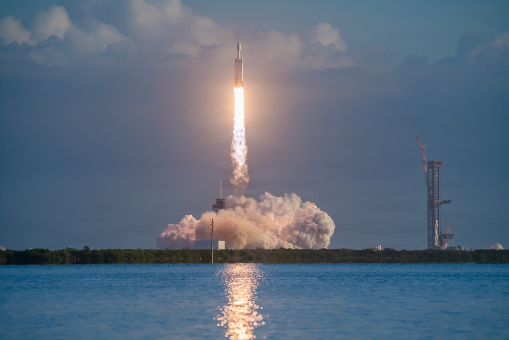 A large rocket is captured mid-launch from a coastal launch pad, with a massive plume of white exhaust smoke billowing outward and a bright flame visible beneath the vehicle. The launch is reflected on the calm water surface in the foreground, suggesting the photographer is positioned across a body of water from the launch site. A secondary launch structure or tower is visible to the right in the background, consistent with a major spaceport facility.
