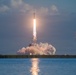 A large rocket is captured mid-launch from a coastal launch pad, with a massive plume of white exhaust smoke billowing outward and a bright flame visible beneath the vehicle. The launch is reflected on the calm water surface in the foreground, suggesting the photographer is positioned across a body of water from the launch site. A secondary launch structure or tower is visible to the right in the background, consistent with a major spaceport facility.