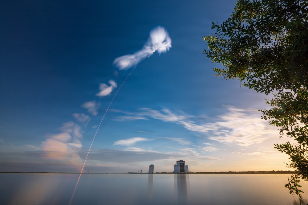 A long-exposure photograph captures a rocket launch trail arcing diagonally across a deep blue sky, originating from a launch facility visible on the horizon. The exhaust plume creates a dramatic white contrail that dissipates and billows at the top of the frame. The scene is reflected in a calm body of water in the foreground, with tree foliage visible on the right side.