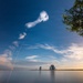 A long-exposure photograph captures a rocket launch trail arcing diagonally across a deep blue sky, originating from a launch facility visible on the horizon. The exhaust plume creates a dramatic white contrail that dissipates and billows at the top of the frame. The scene is reflected in a calm body of water in the foreground, with tree foliage visible on the right side.