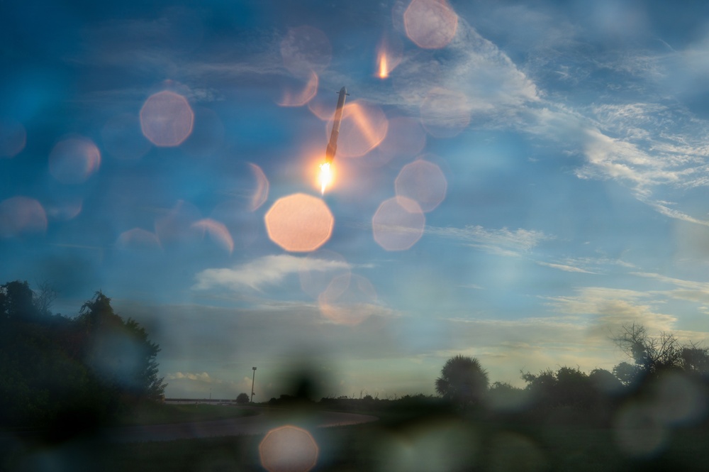A wide-angle photograph taken through a wet or dirty surface, capturing what appears to be a rocket or missile launch against a blue sky with wispy clouds. The image is heavily affected by bokeh circles caused by water droplets or lens distortion on the foreground surface. The scene is set during twilight or early evening, with silhouetted trees and a road visible in the lower portion of the frame.