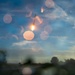 A wide-angle photograph taken through a wet or dirty surface, capturing what appears to be a rocket or missile launch against a blue sky with wispy clouds. The image is heavily affected by bokeh circles caused by water droplets or lens distortion on the foreground surface. The scene is set during twilight or early evening, with silhouetted trees and a road visible in the lower portion of the frame.