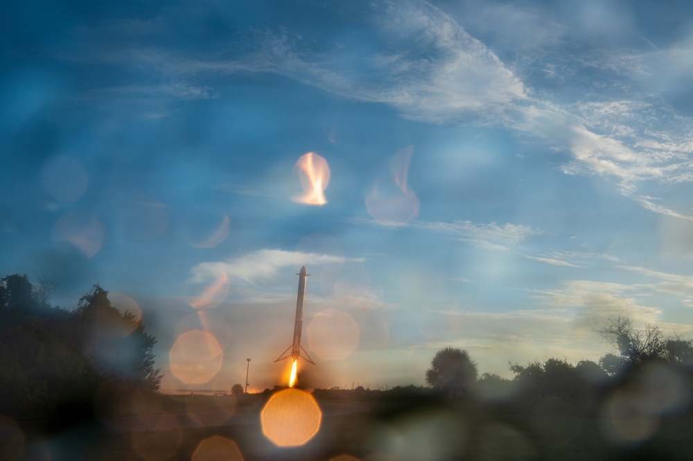 A rocket booster is captured during what appears to be a landing or launch event, with a bright engine flame visible at its base. The image is taken through a wet or rain-speckled surface, creating numerous bokeh circles across the frame. The scene is set against a blue sky with wispy clouds during what appears to be dusk or dawn lighting conditions.