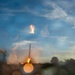 A rocket booster is captured during what appears to be a landing or launch event, with a bright engine flame visible at its base. The image is taken through a wet or rain-speckled surface, creating numerous bokeh circles across the frame. The scene is set against a blue sky with wispy clouds during what appears to be dusk or dawn lighting conditions.