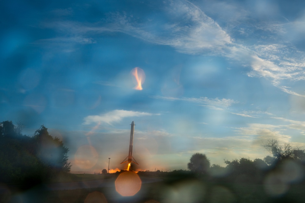 The image captures what appears to be a rocket booster landing or launching at dusk, with visible engine fire and exhaust plume. The scene is photographed through a wet or smudged surface, creating significant lens flare and bokeh effects across the frame. Trees and flat terrain are visible in the background against a blue sky with wispy clouds.
