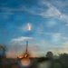 The image captures what appears to be a rocket booster landing or launching at dusk, with visible engine fire and exhaust plume. The scene is photographed through a wet or smudged surface, creating significant lens flare and bokeh effects across the frame. Trees and flat terrain are visible in the background against a blue sky with wispy clouds.