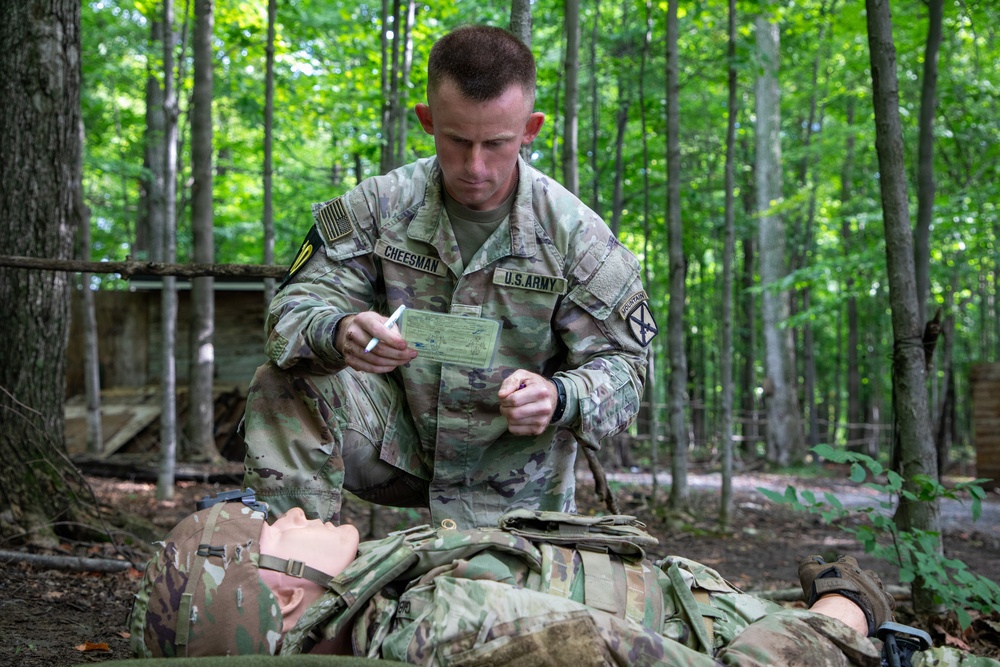 A U.S. Army soldier identified by name tape as 'CHEESMAN' is kneeling over a simulated casualty (mannequin) in a wooded training environment. He appears to be filling out or reviewing a field medical card, consistent with combat casualty care training. The setting suggests a tactical combat casualty care (TCCC) or field training exercise scenario.