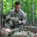 A U.S. Army soldier identified by name tape as 'CHEESMAN' is kneeling over a simulated casualty (mannequin) in a wooded training environment. He appears to be filling out or reviewing a field medical card, consistent with combat casualty care training. The setting suggests a tactical combat casualty care (TCCC) or field training exercise scenario.