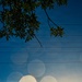 An outdoor photograph taken looking upward toward a blue sky during golden hour or sunset, with tree branches and leaves visible in the upper left corner. The image features prominent lens flare artifacts appearing as multiple large octagonal bokeh shapes caused by direct sunlight entering the camera lens. A silhouetted structure, possibly a utility pole or antenna, is visible near the bottom center against the bright horizon.
