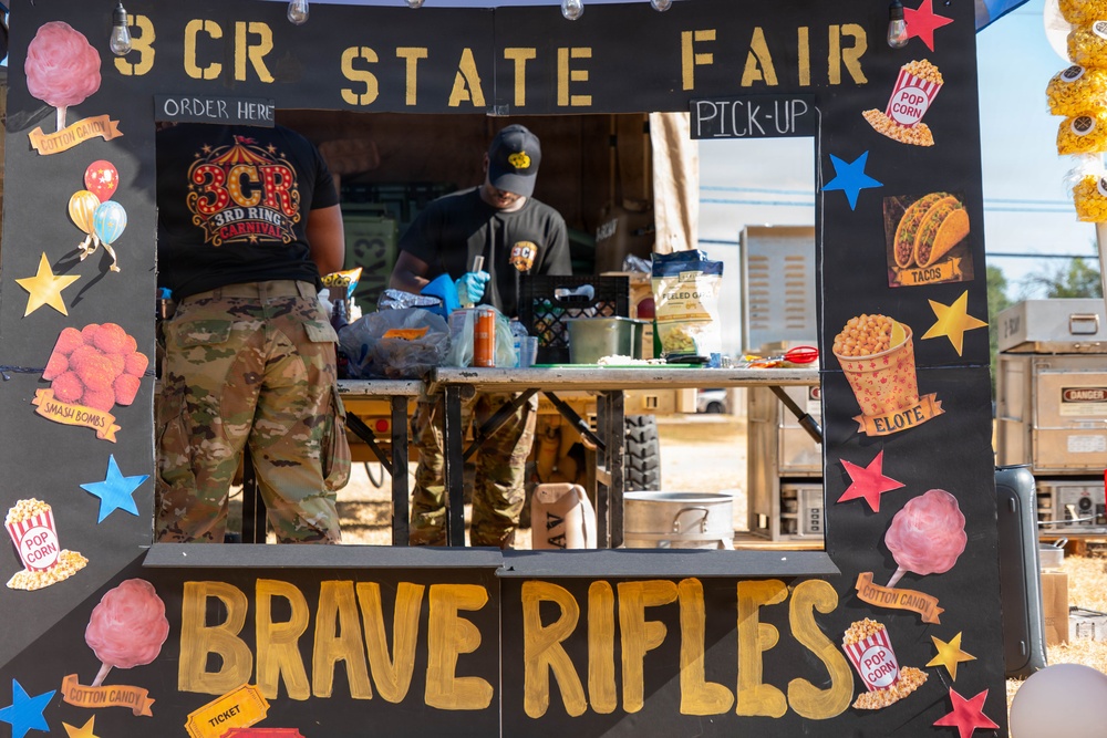 The image shows a military-themed state fair food booth labeled '3CR State Fair' and 'Brave Rifles,' operated by soldiers in camouflage uniforms. The booth is decorated with colorful illustrations of fair foods including cotton candy, popcorn, tacos, elote, and smash bombs. Two service members are visible working behind the counter, which is framed by a large decorative black board with gold lettering.