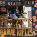 The image shows a military-themed state fair food booth labeled '3CR State Fair' and 'Brave Rifles,' operated by soldiers in camouflage uniforms. The booth is decorated with colorful illustrations of fair foods including cotton candy, popcorn, tacos, elote, and smash bombs. Two service members are visible working behind the counter, which is framed by a large decorative black board with gold lettering.