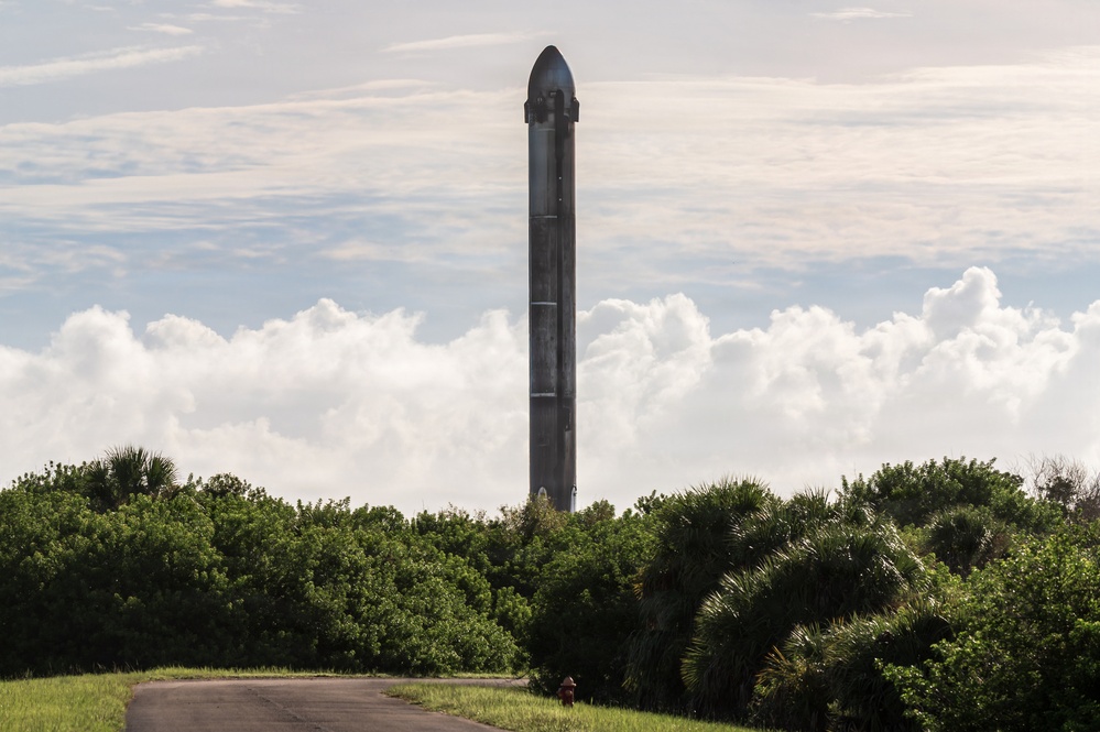 A tall, dark-colored rocket booster stands vertically on a launch pad, visible above a treeline of dense green vegetation including palm trees. The structure appears to be a reusable rocket booster, likely a SpaceX Falcon 9 or similar vehicle, photographed from a distance along a paved road. The scene is set against a partly cloudy sky with large cumulus clouds in the background.