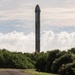 A tall, dark-colored rocket booster stands vertically on a launch pad, visible above a treeline of dense green vegetation including palm trees. The structure appears to be a reusable rocket booster, likely a SpaceX Falcon 9 or similar vehicle, photographed from a distance along a paved road. The scene is set against a partly cloudy sky with large cumulus clouds in the background.