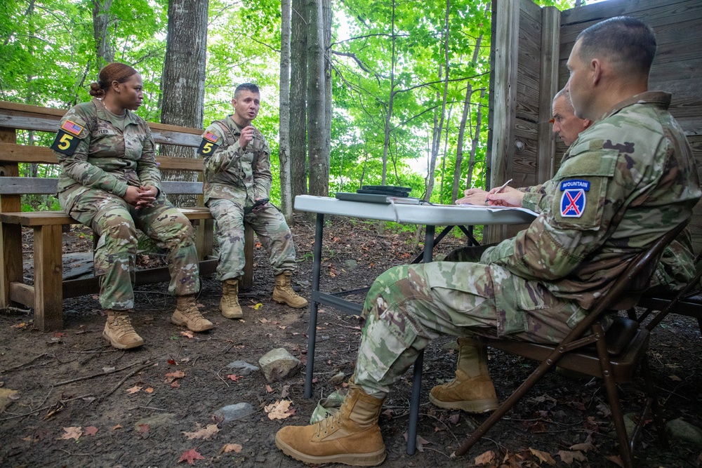 Four U.S. Army soldiers in OCP (Operational Camouflage Pattern) uniforms are engaged in what appears to be a formal evaluation or debriefing session in an outdoor wooded setting. Two soldiers seated on a bench, both wearing '5' numbered patches, face two evaluators seated at a folding table. The soldier on the right side of the table wears a 10th Mountain Division patch, suggesting this may be a training assessment or after-action review.